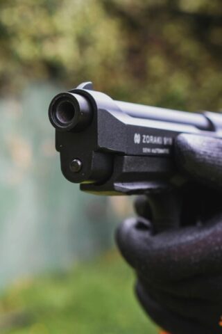 A detailed close-up of a gloved hand holding and aiming a semi-automatic pistol outdoors.
